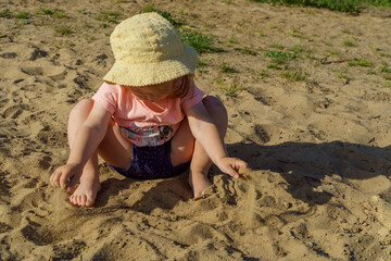 little girl on the beach in a yellow hat having fun playing with sand girl wearing a pink t-shirt and blue shorts girl very happy and interested in sunny warm weather in summer on vacation