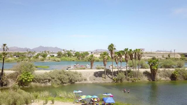aerial footage of the silky green waters of the Colorado River with people in boats on the water surrounded by lush green palm trees and grass with blue sky at Quechan Park in Blythe California USA