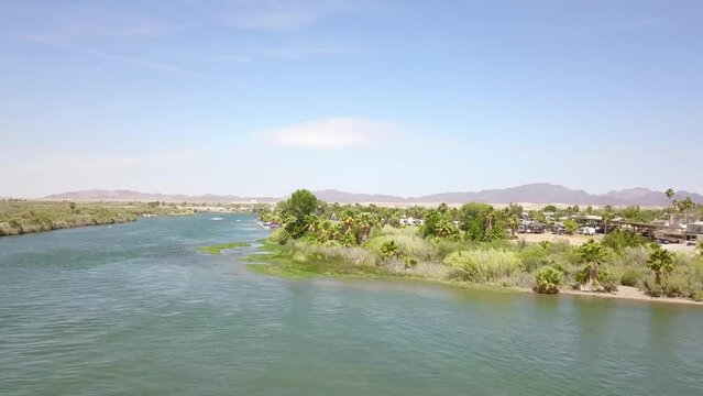 aerial footage of the silky green waters of the Colorado River with people in boats on the water surrounded by lush green palm trees and grass with blue sky at Quechan Park in Blythe California USA