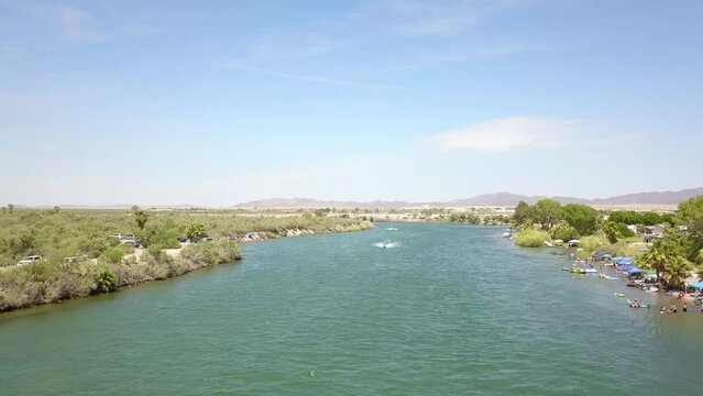 aerial footage of the silky green waters of the Colorado River with people riding jet skis and lush green trees and plants on the banks of the river with blue sky and clouds at Quechan Park in Blythe 