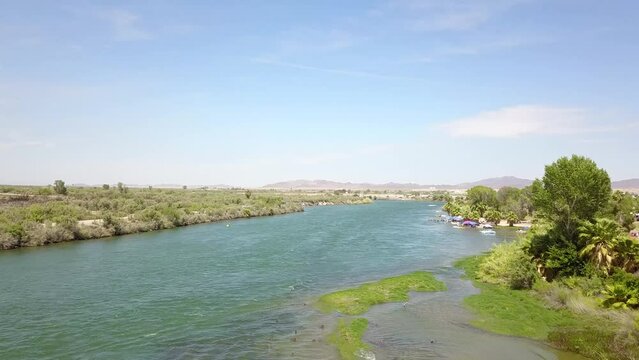 aerial footage of the silky green waters of the Colorado River with people riding jet skis and lush green trees and plants on the banks of the river with blue sky and clouds at Quechan Park in Blythe 