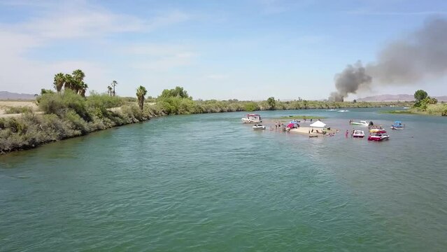 aerial footage of the green waters of the Colorado River with people in boats and smoke from a brush fire on the banks of the river with blue sky at Quechan Park in Blythe California USA
