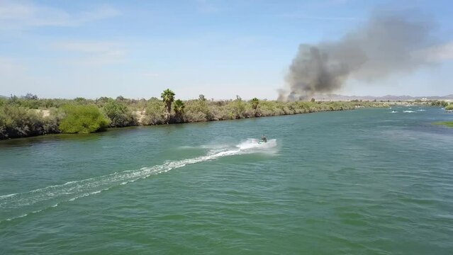aerial footage of the green waters of the Colorado River with people in boats and smoke from a brush fire on the banks of the river with blue sky at Quechan Park in Blythe California USA
