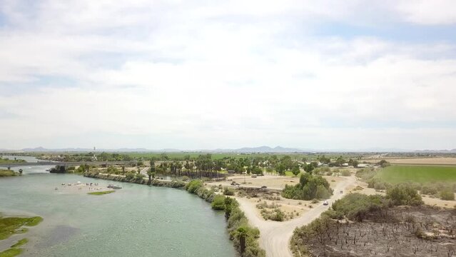 aerial footage of the silky green waters of the Colorado river with people riding jet skis and sailing boats and a street with cars driving and lush green palm trees and grass at Quechan Park