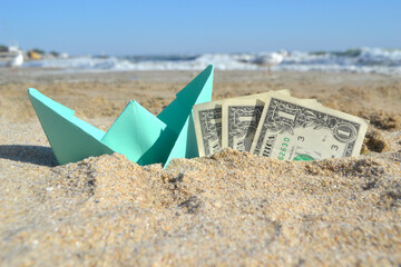 Blue small paper boat and three dollar paper bills half in sand on beach near sea on summer sunny day. Concept of travel, dreams, expenses, money, finance, tourism, rest, vacation holiday goals wishes