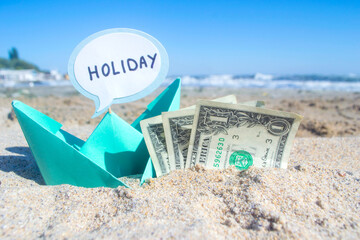 Small blue paper boat, three one paper dollar bills and speech bubble with word Holiday on sandy sea beach on sunny summer day. Concept money finance business tourism travel dreams vacation holiday