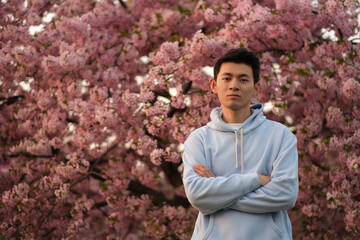 cool handsome Asian young man arm crossing, looking at camera. Blur pink sakura tree background
