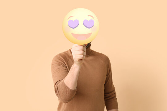 Young man holding happy emoticon with heart-shaped eyes on color background