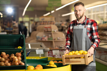 Close up portrait of joyful male supermarket worker in good mood holding in hands box with fresh organic fruits, looking at camera and smiling, arranging citrus fruits in the fruit department