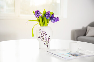 Stylish vase with hyacinth flowers, newspaper and cup of coffee on table in room