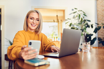 Cheerful mature woman wearing glasses using smartphone working at home office