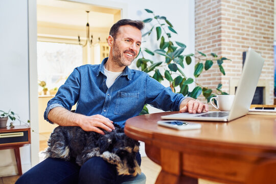 Handsome mid adult man using laptop sitting at a table at home with dog sleeping on lap