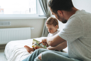 Happy father young man and baby girl little daughter having fun reading a book in children room at...