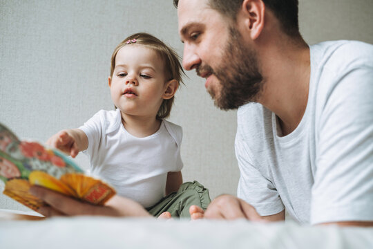 Happy father young man and baby girl little daughter having fun reading a book in children room at home