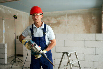 Fototapeta premium Professional construction worker in uniform standing with rotary hammer drill. Portrait of contractor in hardhat and overalls posing with jackhammer near step ladder and masonry indoors.