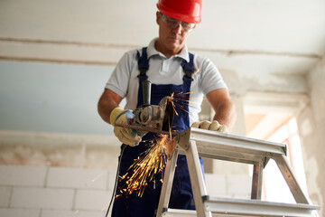 Professional construction worker hands in work gloves using angle grinder to cut metal rod at building site. Close up shot of contractor cutting iron stick on ladder with glowing sparks.