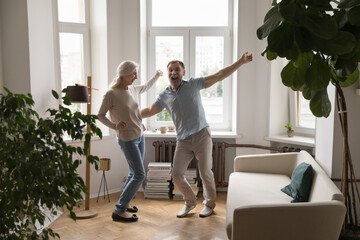 Excited happy senior couple dancing to music together at home couch, enjoying date, anniversary, active entertainment, party for two, having fun together. Retirement, indoor activity concept - obrazy, fototapety, plakaty
