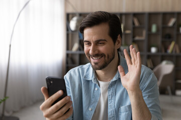 Hello. Happy millennial man wave hand hi look at smartphone screen start talk by video call with...
