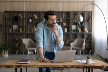 Work on idea. Thoughtful casual male in glasses stand by desk with pc look at distance touch chin...