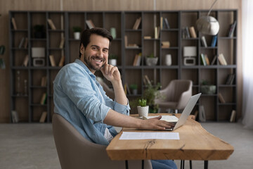 Young worker. Portrait of confident man specialist work at modern home office study in library using laptop computer. Smiling male expert sit at workplace desk look at camera distracted from pc screen
