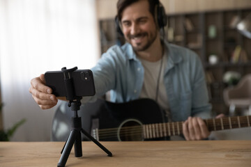 Smiling young man artist in headphones prepare internet stream from home domestic studio hold guitar set up phone app. Focus on male singer hand putting smartphone on holder starting live broadcast