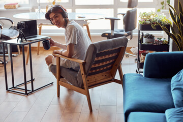 Smiling Asian guy holding cup while working with laptop at home