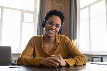 Smiling African woman in headphones looks at camera take part in virtual meeting by study or business using video conference application. Modern tech, remote communication, video call event concept