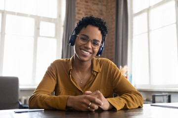 Smiling African woman in headphones looks at camera take part in virtual meeting by study or...