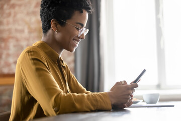 Side profile view young smiling African woman in glasses sit at table use smartphone. Share text message, take break at workplace spend time on internet, scroll news feed in social media, Tech concept - obrazy, fototapety, plakaty