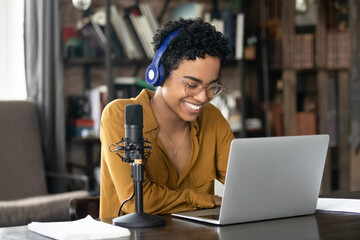 Smiling young African woman podcaster in headphones sit at desk in front of laptop and microphone....