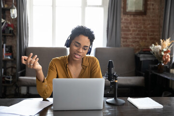 African woman wear wireless headphones sit at desk looks at laptop screen make speech talk into...