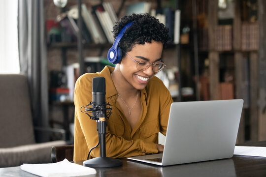 Smiling young African woman podcaster in headphones sit at desk in front of laptop and microphone. Share information, make audio podcast for internet audience, lead on-line stream, blogging concept