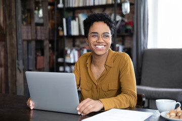 Happy African woman freelancer distracted from working on laptop smile staring into distance, sit at workplace desk studying online, take remote professional training use modern tech, telework concept
