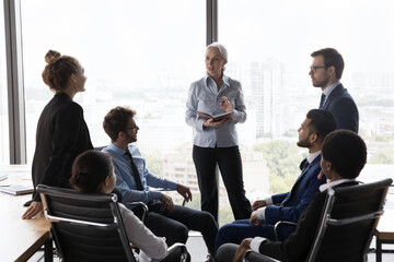 Older female company boss makes speech to multi ethnic staff members, leads morning briefing in modern office boardroom. Leadership, seminar corporate training, share experience to youngers concept