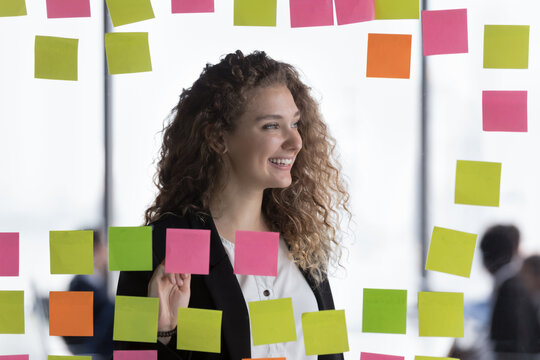 Young attractive woman pose in modern office look into distance, view through transparent wall with attached sticky notes post-it reminders. Creative approach to solve business tasks, planning concept
