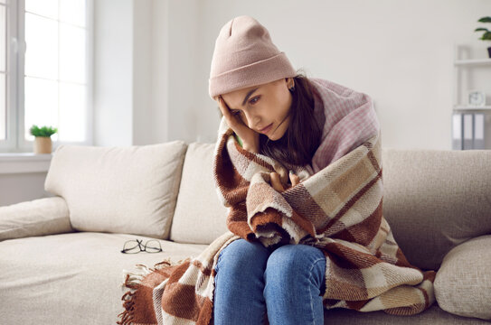It's cold in the house in winter. Woman freezing because of fever or trouble with central heating. Sad lady wrapped in wool plaid and scarf and wearing warm hat sitting on sofa at home in wintertime