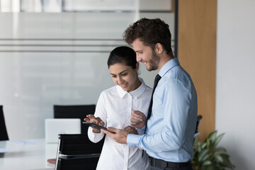 Indian woman with male colleague use digital tablet standing in office, review new business application on device, discuss collaborative project, engaged in learn online task during break at workplace