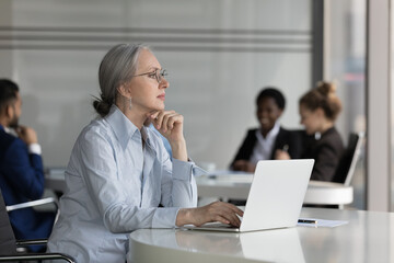 Thoughtful middle-aged businesswoman sit at desk with laptop looks into distance, search business...