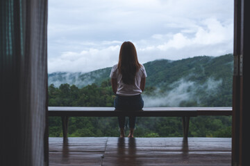 Rear view image of a woman sitting on wooden balcony while watching a beautiful mountains and nature view on foggy day