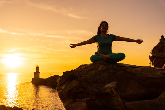 A woman doing meditation and yoga exercises at sunset next to a lighthouse in the sea, healthy and naturist life, outdoor pilates, ardha padmasana