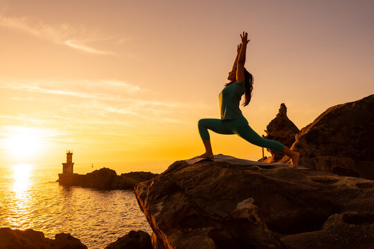 Anjaneyasana, a woman doing meditation and yoga exercises on a rock at sunset next to a lighthouse in the sea, healthy and naturist life