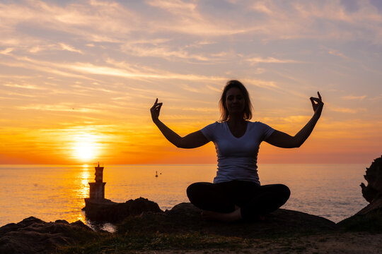 A blonde woman doing meditation on a rock at sunset next to a lighthouse in the sea, healthy and naturist life