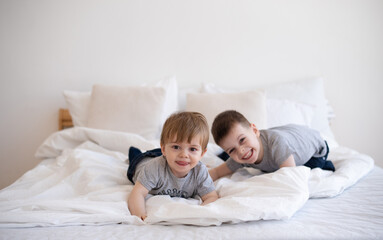 Fototapeta na wymiar Smiling two boys in gray t-shirts lie and playing on white blanket on bed. Cheerful and happy childhood in family. Exercise and play good for health. Together joyful and interesting. 