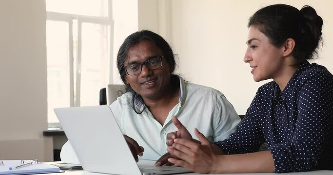 Friendly young woman agent broker designer hold dialogue with man client discuss project vision listen to customer propositions. Indian business partners converse by laptop cooperate in job on startup