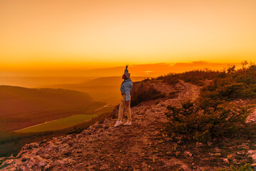 Fototapeta premium Woman tourist on top of sunrise mountain. The girl salutes the sun, wearing a blue jacket, white hat and white jeans. Conceptual design.