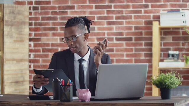Office black man with dreadlocks and glasses with suit speaks on the phone, sitting at a desk a laptop and takes paper tablet in hand.