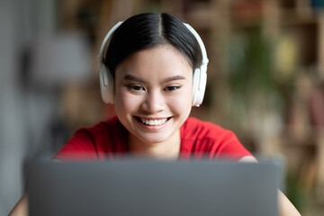 Sad tired young chinese girl student in wireless headphones lies on table, looks at laptop in room