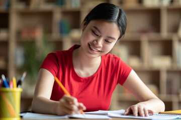 Cheerful pretty smart young chinese girl student making notes, homework, studying in living room