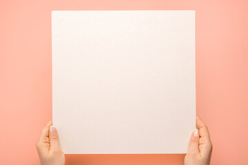 Obraz premium Mockup square paper. Overhead shot of woman hands with blank paper sheet on pink table. Human hands holding mockup white advertising card. Isolated on pastel background