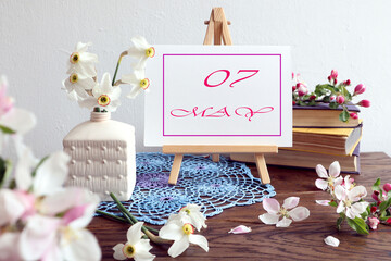 Calendar for May 7 : an easel with an inscription - the name of the month of May in English, the numbers 07, a bouquet of daffodils in a white vase, apple branches on a blue napkin, a stack of books